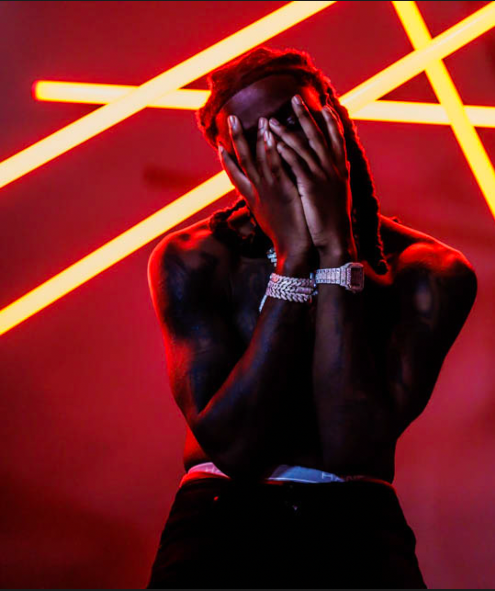 A shirtless Sleazy World Go covers his face with hands, wearing diamond bracelets, posed against red neon lights.