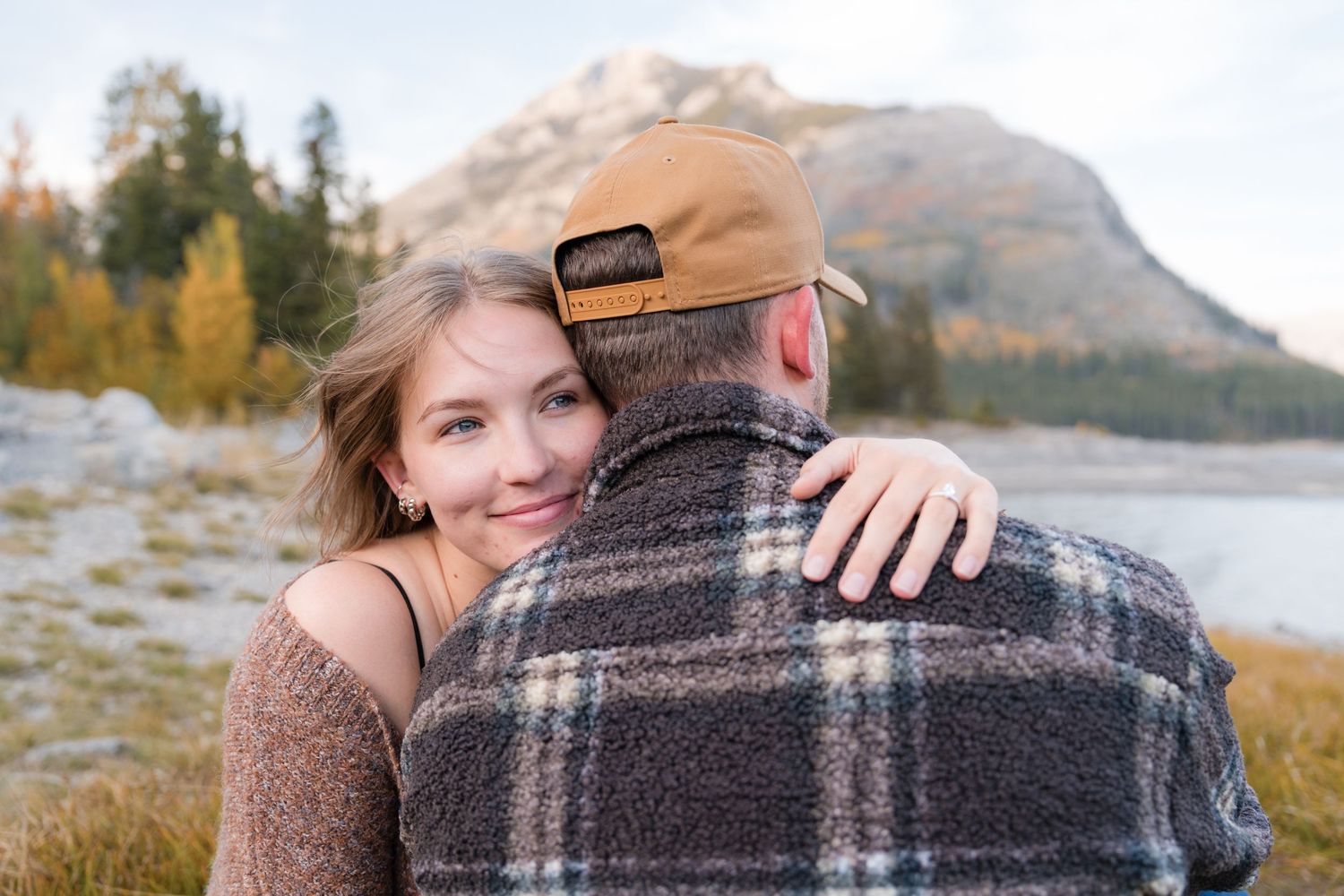 Couple embracing at Lake Minnewanka, Banff in autumn, woman smiling over man's shoulder wearing plaid jacket.