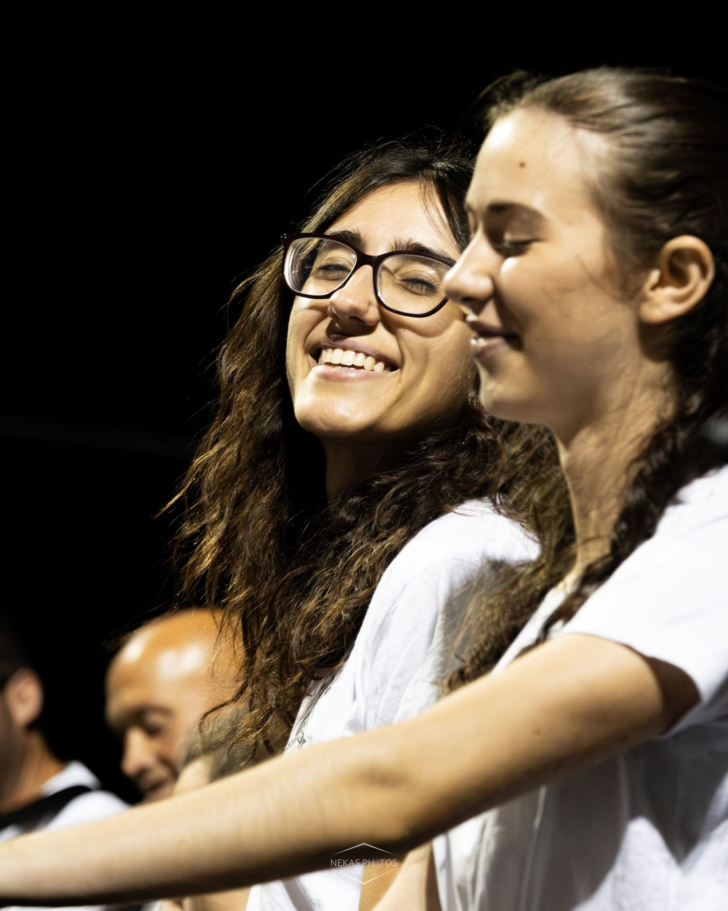 Two young women smiling and laughing together at an outdoor event, one wearing glasses and a white shirt.
