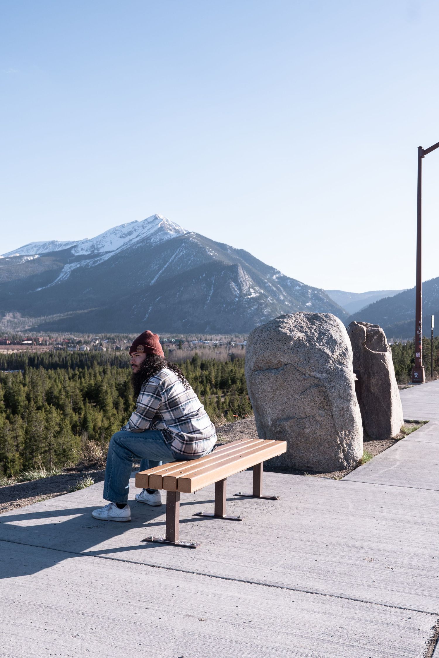 Person sitting on a wooden bench at a mountain overlook with snow-capped peaks and forest views on a clear day.