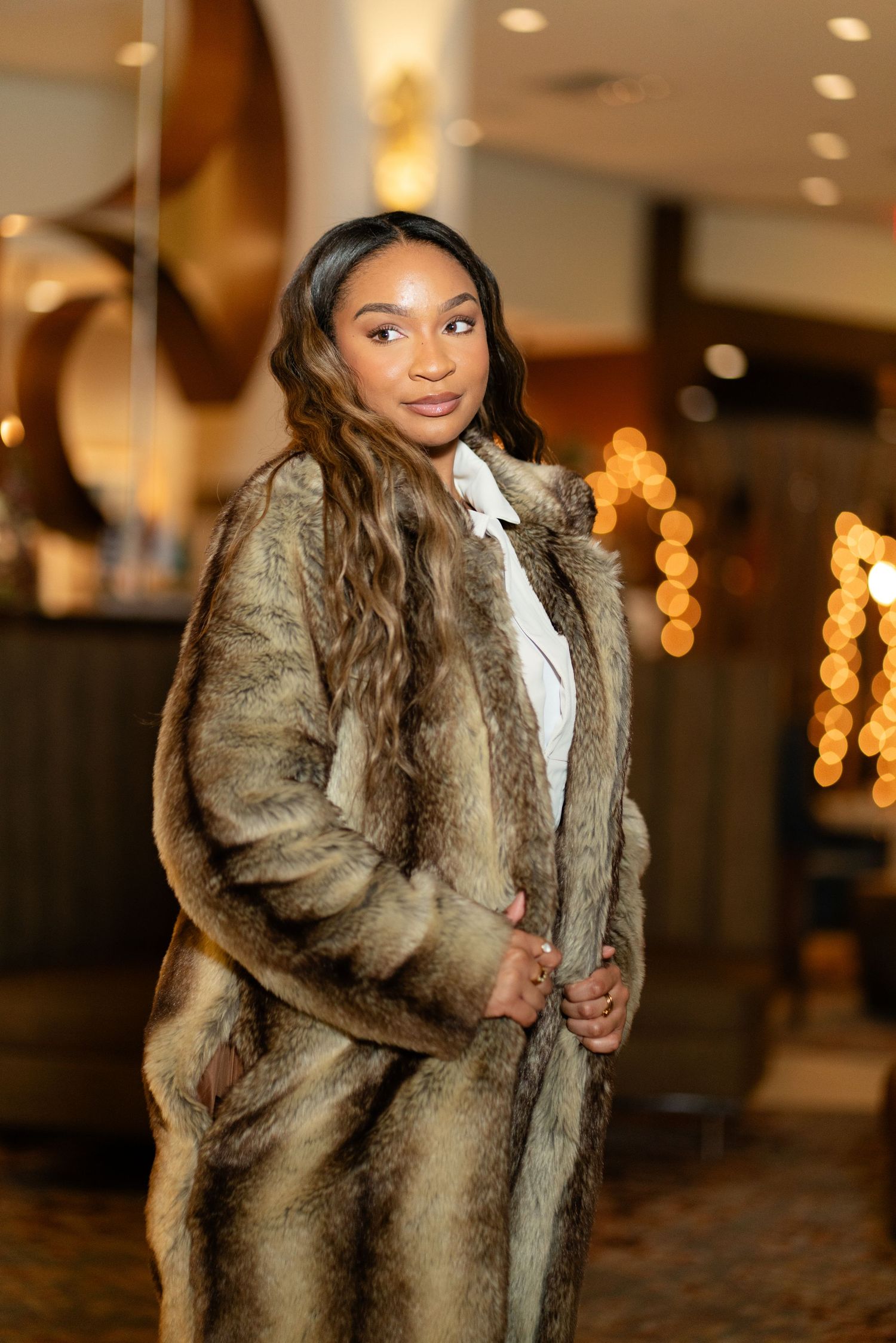Model poses in a luxurious brown fur coat against a warmly lit indoor backdrop with bokeh lights.