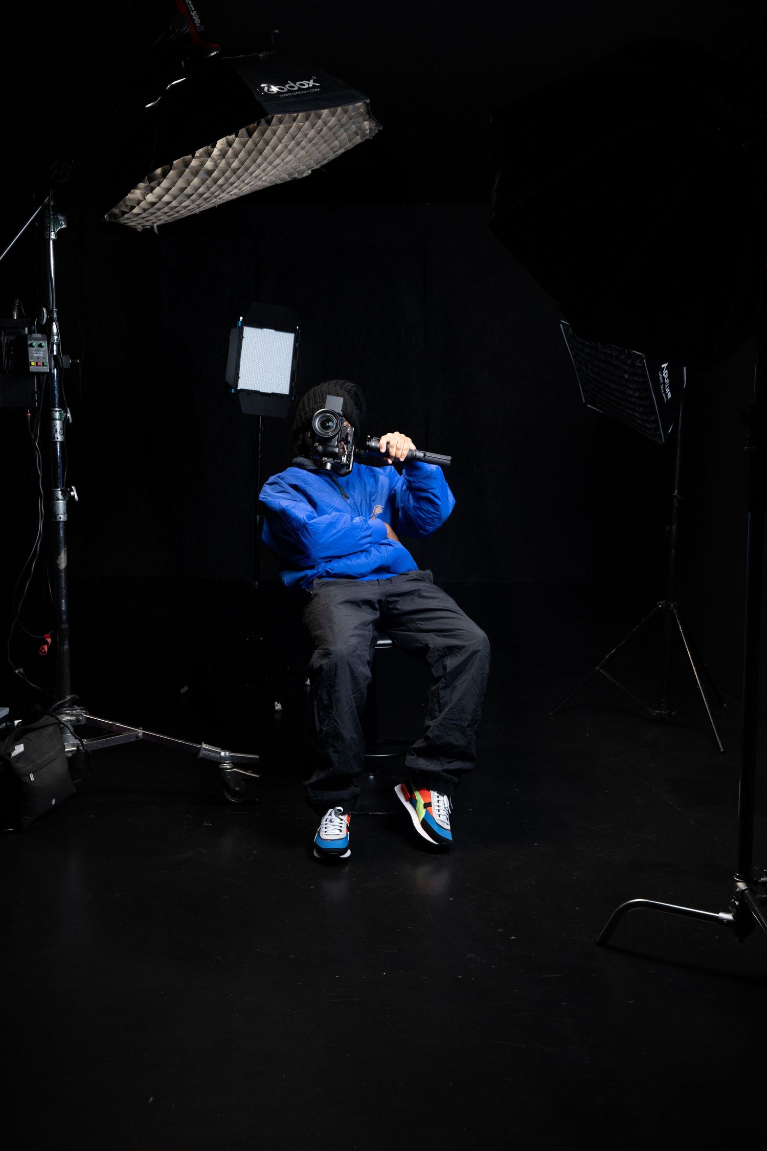 A person wearing blue and black clothing sits in a dark studio setting with dramatic lighting and photographic equipment.