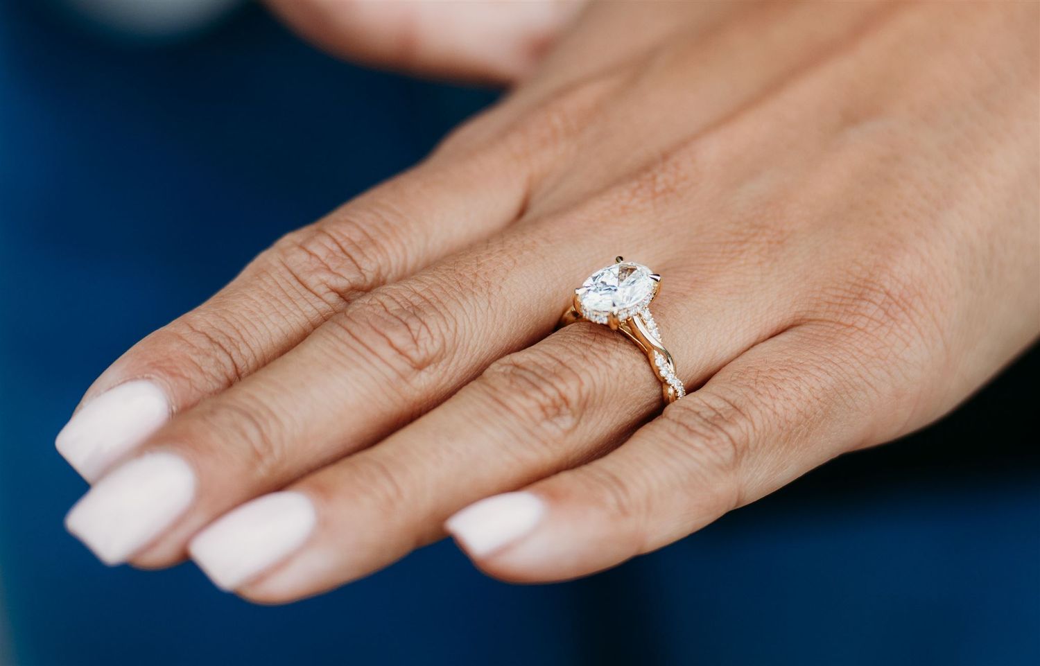 Close-up of a hand with white nails wearing a gold engagement ring featuring a large oval diamond solitaire.