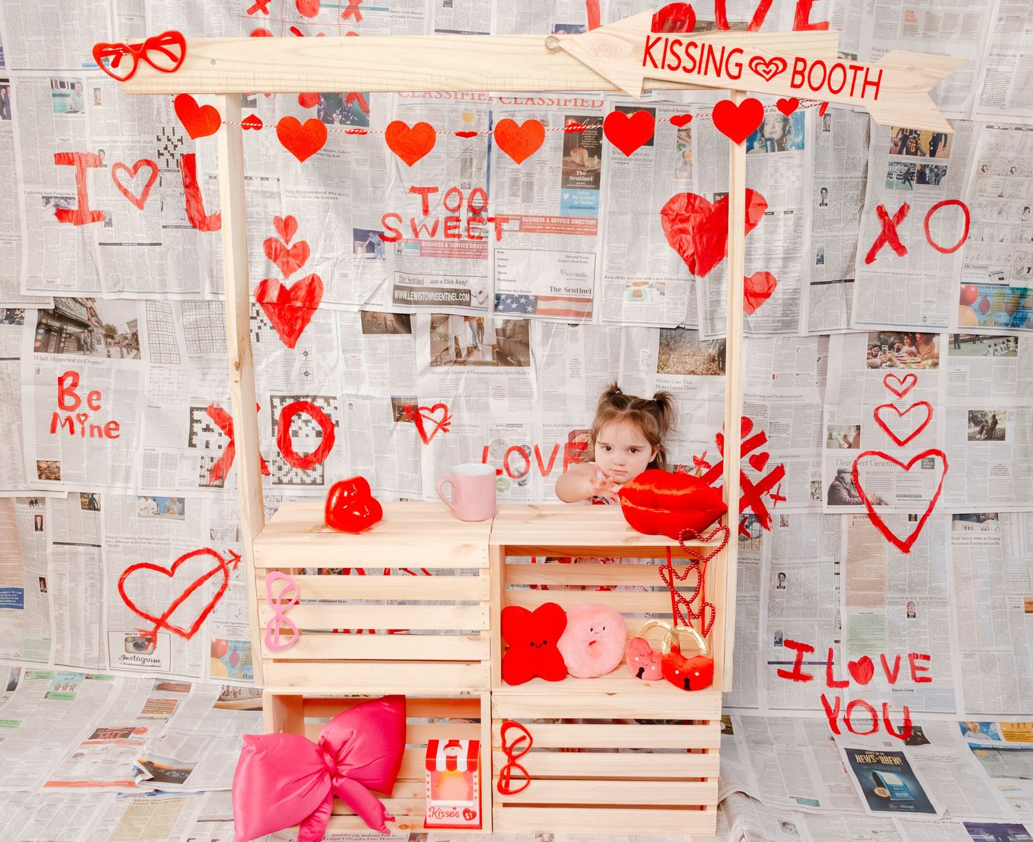 Baby in white dress with glasses sits in wooden crate beside FRAGILE box and lamp, Christmas tree background.