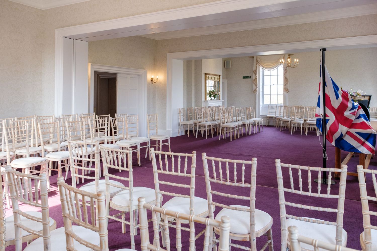 Elegant wedding ceremony room with white Chiavari chairs, purple carpet, and a Union Jack flag in a bright, formal venue.