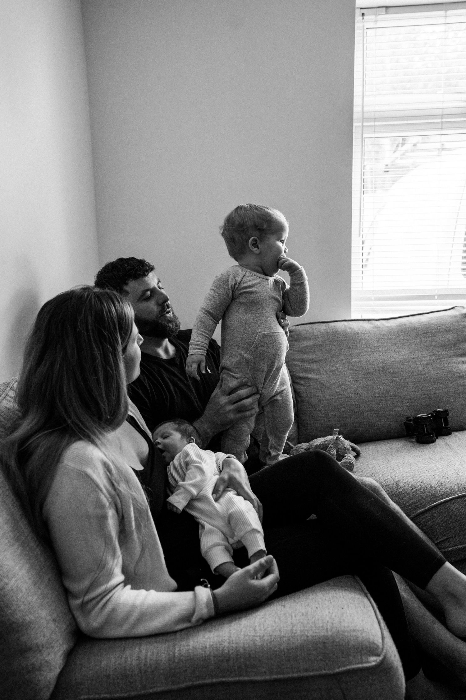 Black and white photo of a family relaxing on a couch, with a baby on mom's lap and a toddler standing beside dad.