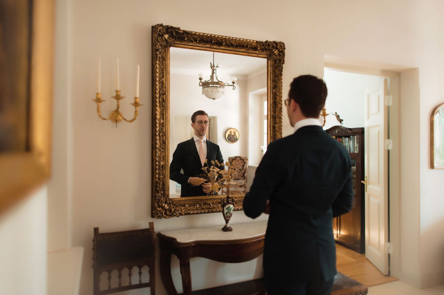 Groom in dark suit adjusts tie while viewing reflection in ornate gold-framed mirror in elegant room.