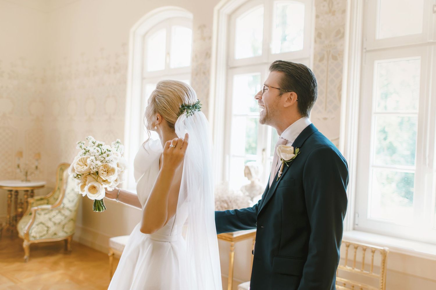 Bride and groom share a tender moment by elegant windows, bride holding white bouquet in a classic wedding venue.