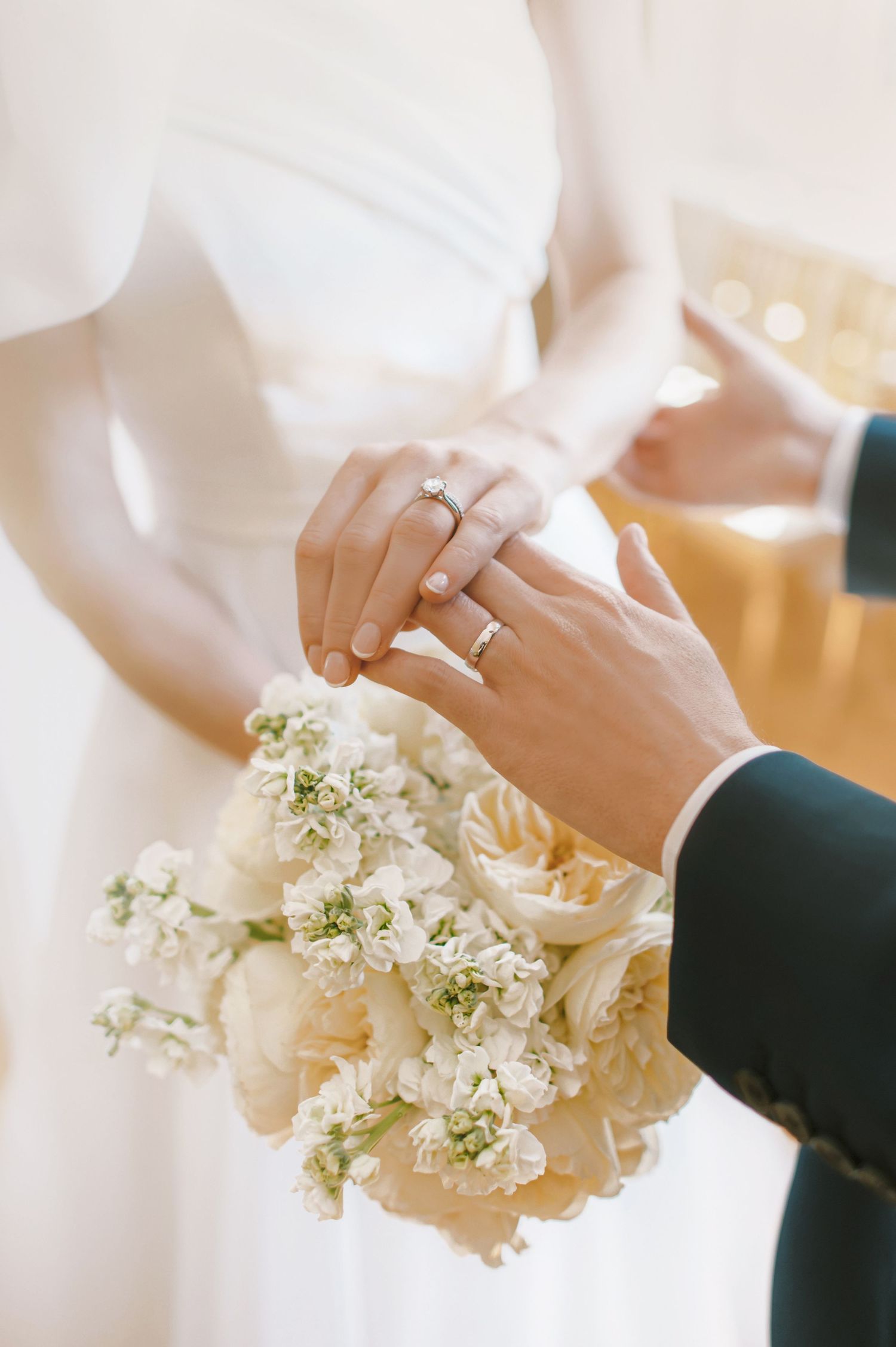 Bride and groom exchanging rings during wedding ceremony, holding white floral bouquet with roses and delicate blooms.