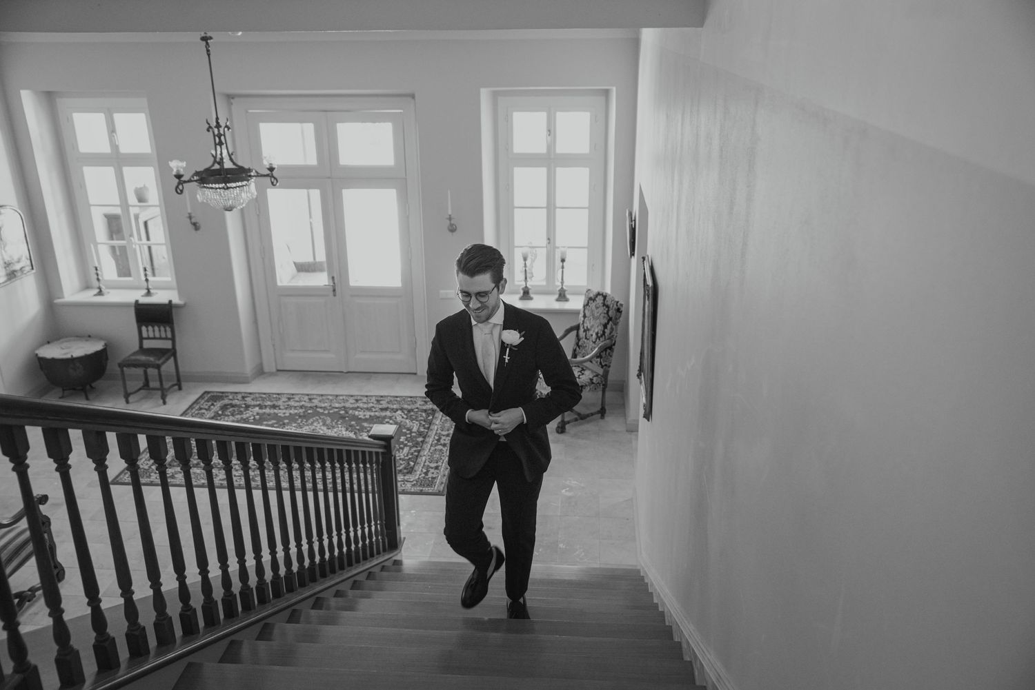 Groom in black suit descending elegant staircase in black and white wedding photo.