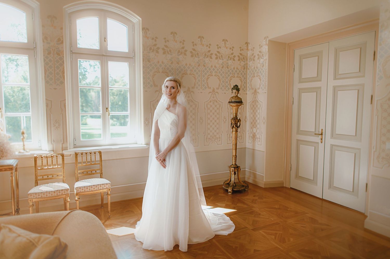 Bride in white gown and veil stands in elegant room with ornate walls, parquet floors, and large windows.