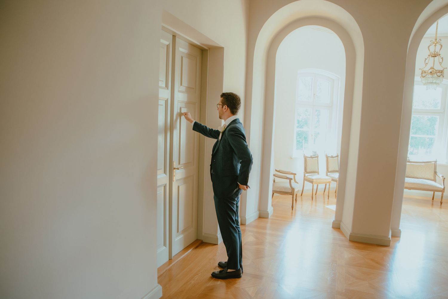 Groom in dark suit standing in elegant hallway with arched doorways and hardwood floors.