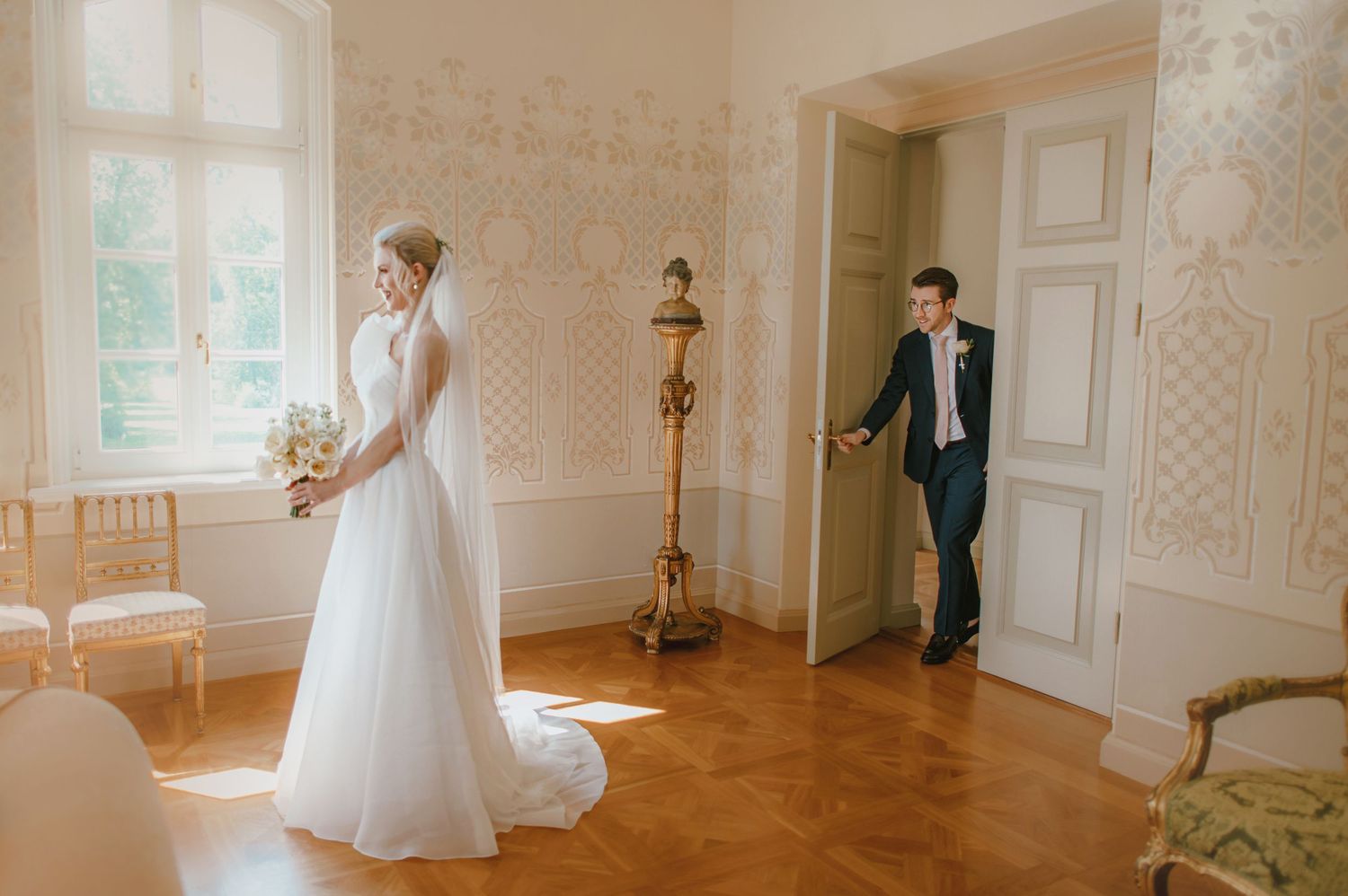Bride in white gown holds bouquet as groom enters elegant room during wedding first look ceremony.