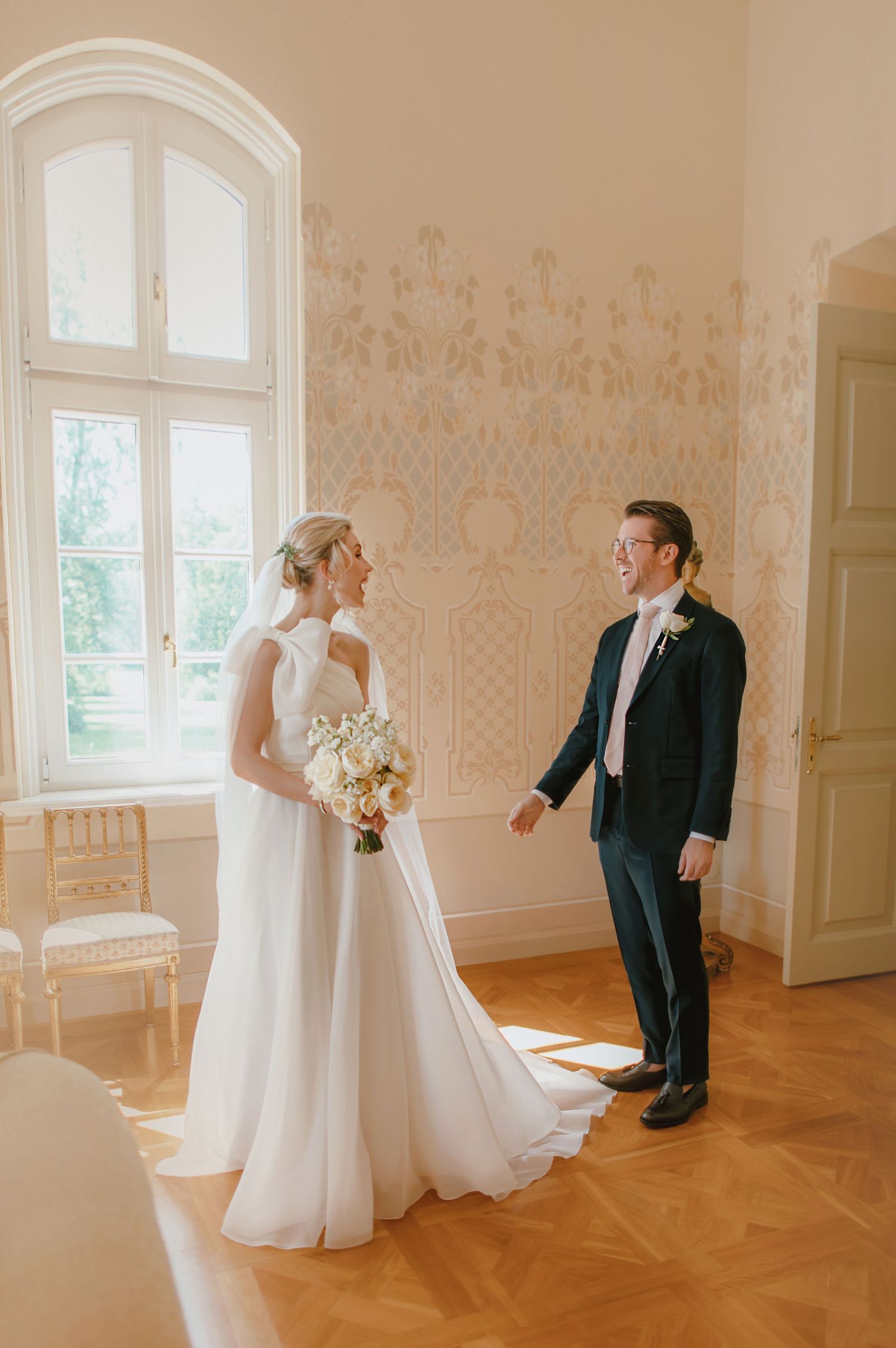 Bride in white gown holds bouquet as groom in navy suit smiles during first look in elegant room.