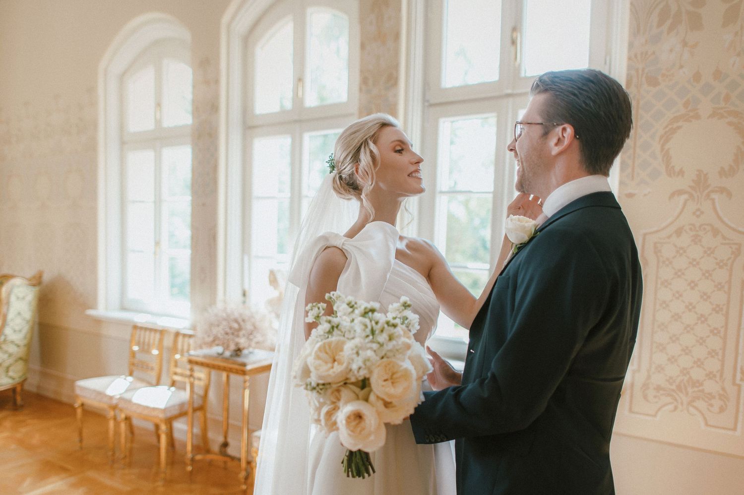 Bride and groom share a joyful moment indoors, she holds white roses bouquet, he adjusts tie in elegant room.