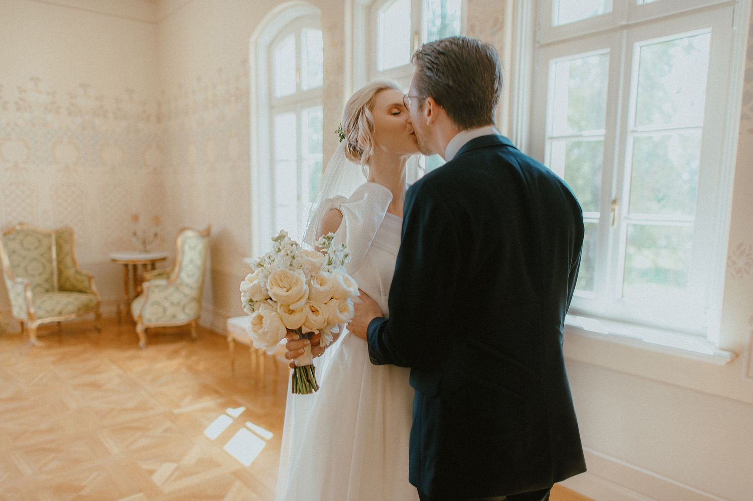 Bride and groom sharing a first kiss at their wedding ceremony in an elegant sunlit room with large windows.