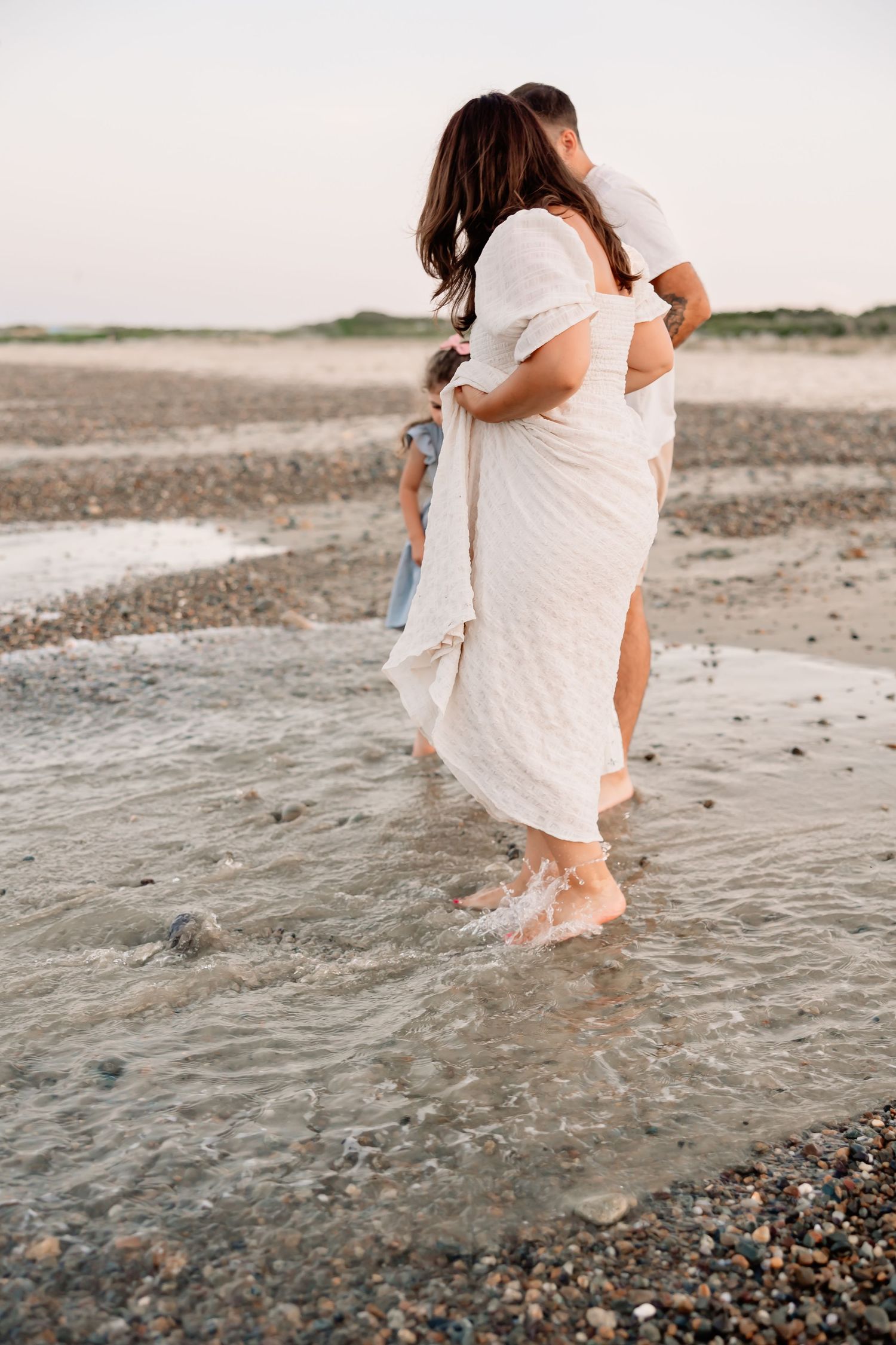 A couple embraces barefoot in shallow beach water, woman in white dress, child nearby on rocky shore at sunset.