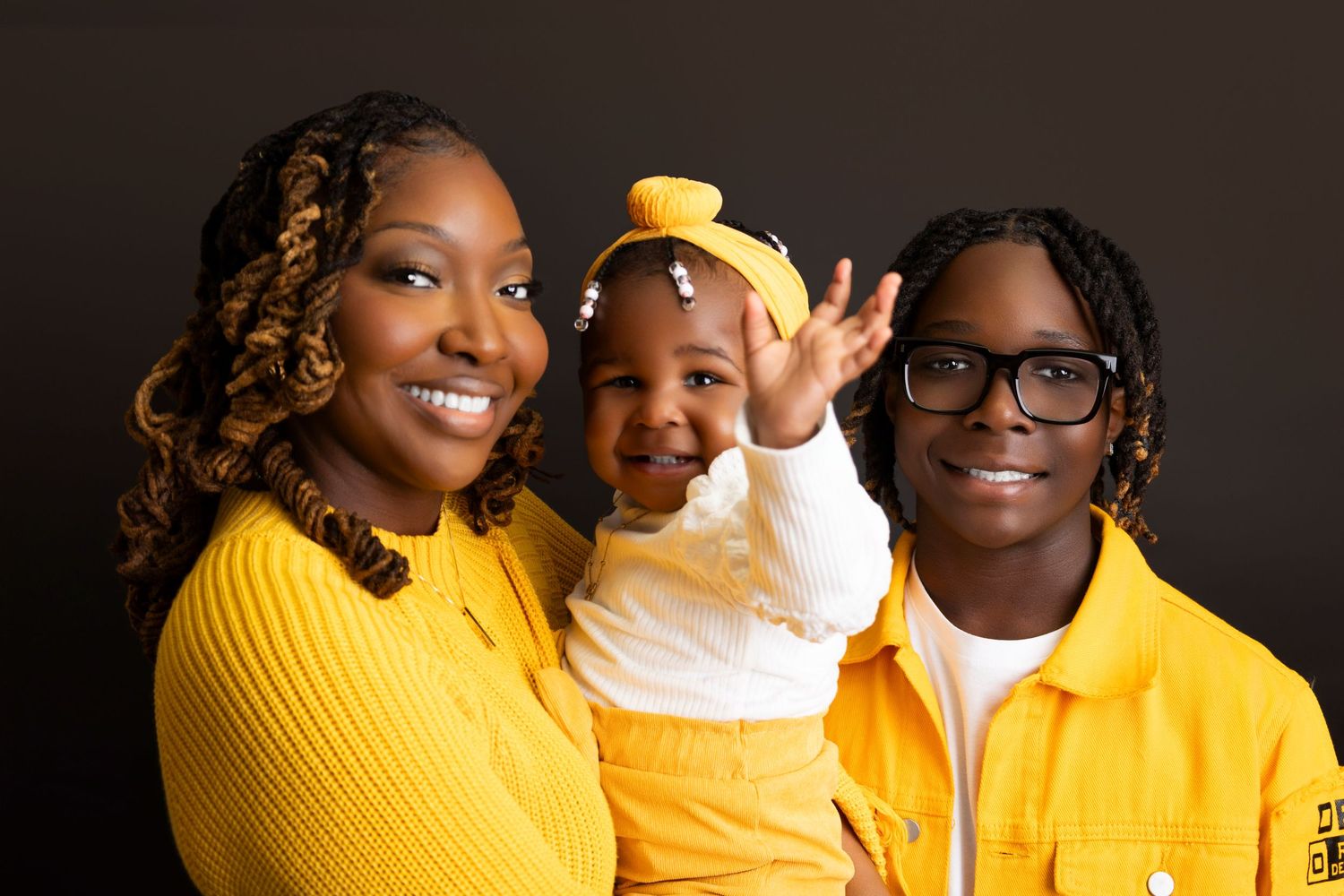 Family portrait of two adults and a baby in matching yellow outfits smiling against a dark background.