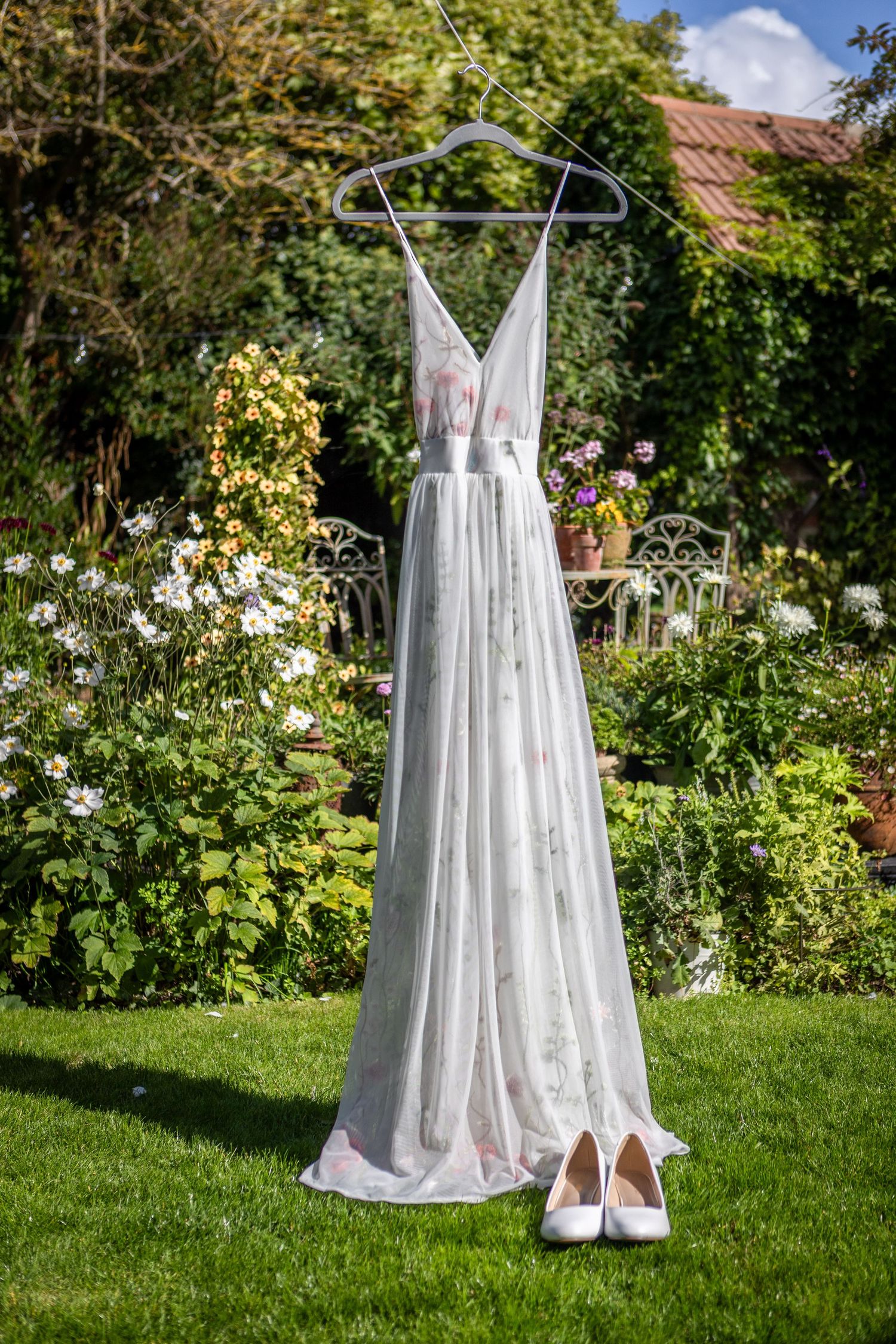 Elegant white floral wedding dress hangs on hanger above white heels in a sunny blooming garden.