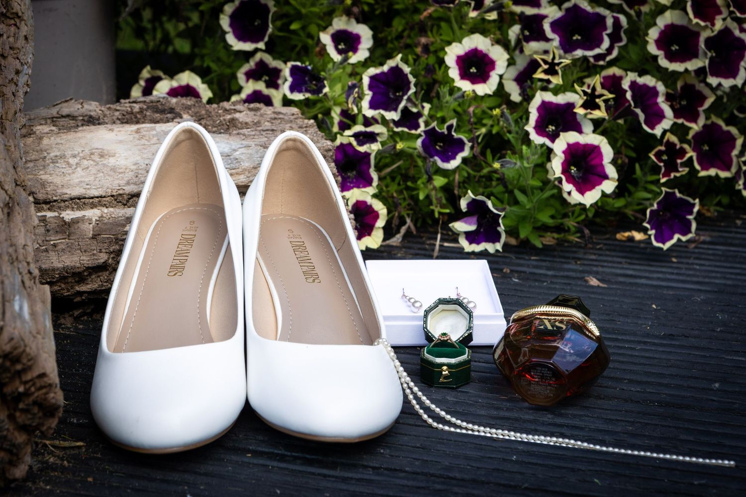 White bridal heels displayed with wedding ring and accessories near purple petunia flowers.