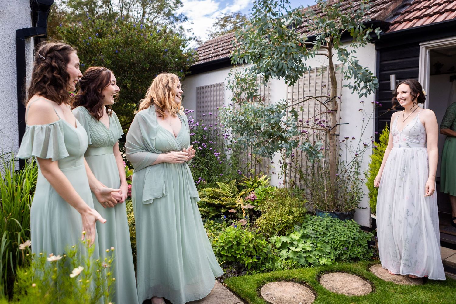 Bride in flowy grey gown surprises three bridesmaids in mint dresses in a charming cottage garden.