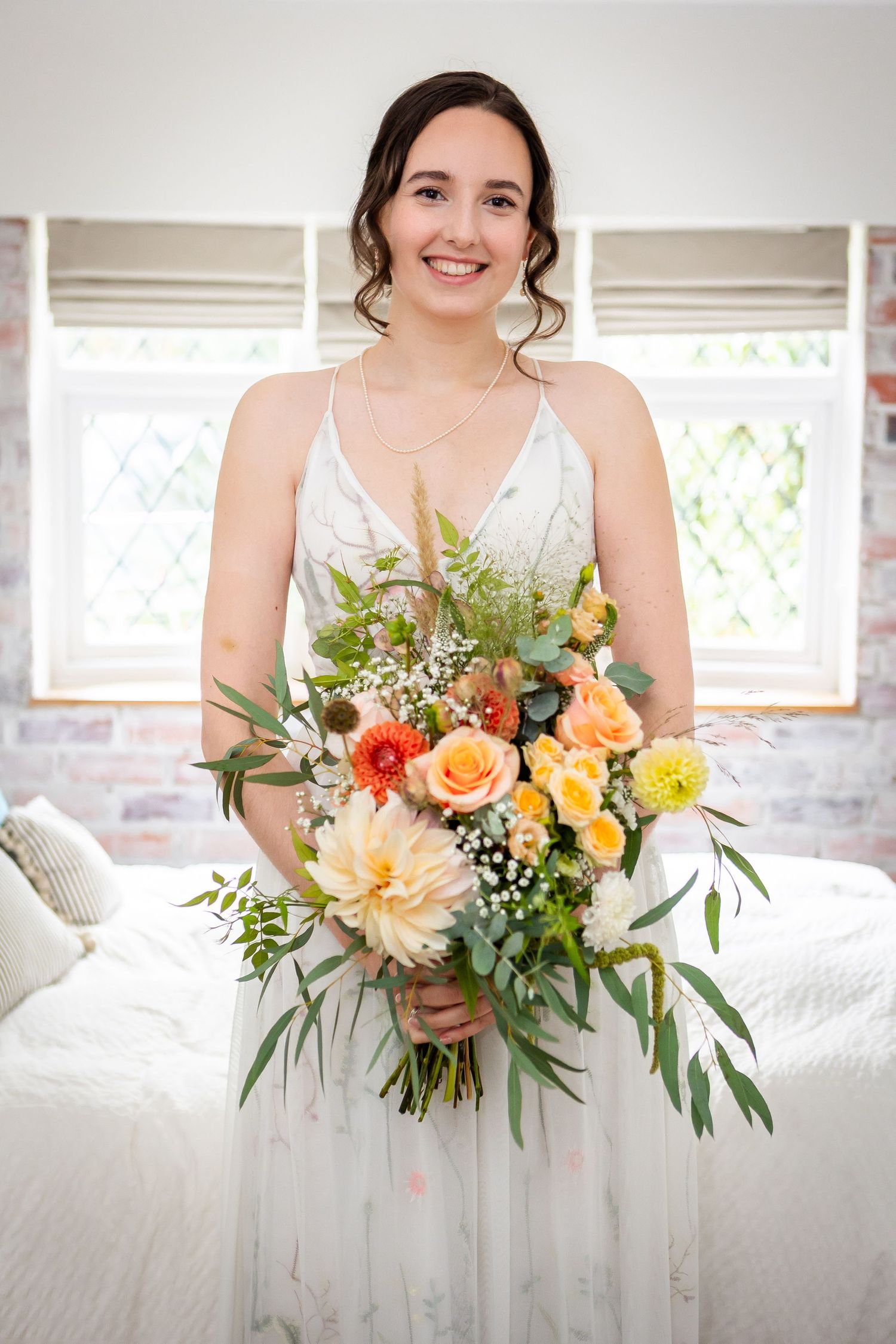 Smiling bride in white dress holding colorful bouquet of orange roses and dahlias in a bright bedroom.