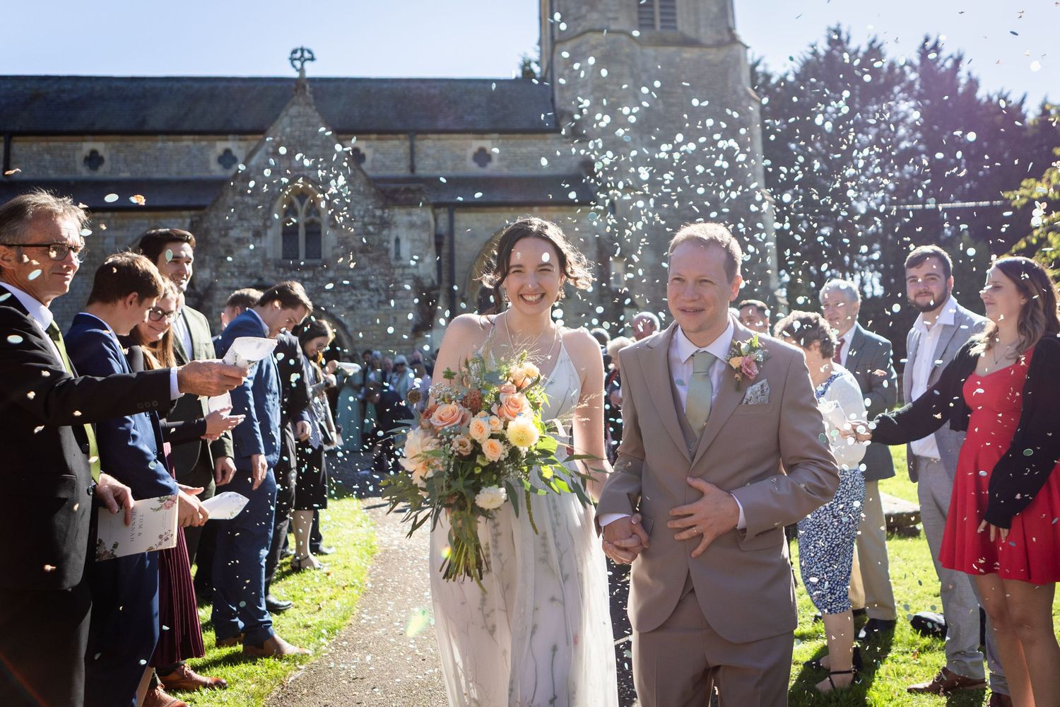 Bride and groom walk through confetti shower outside a stone church surrounded by wedding guests.