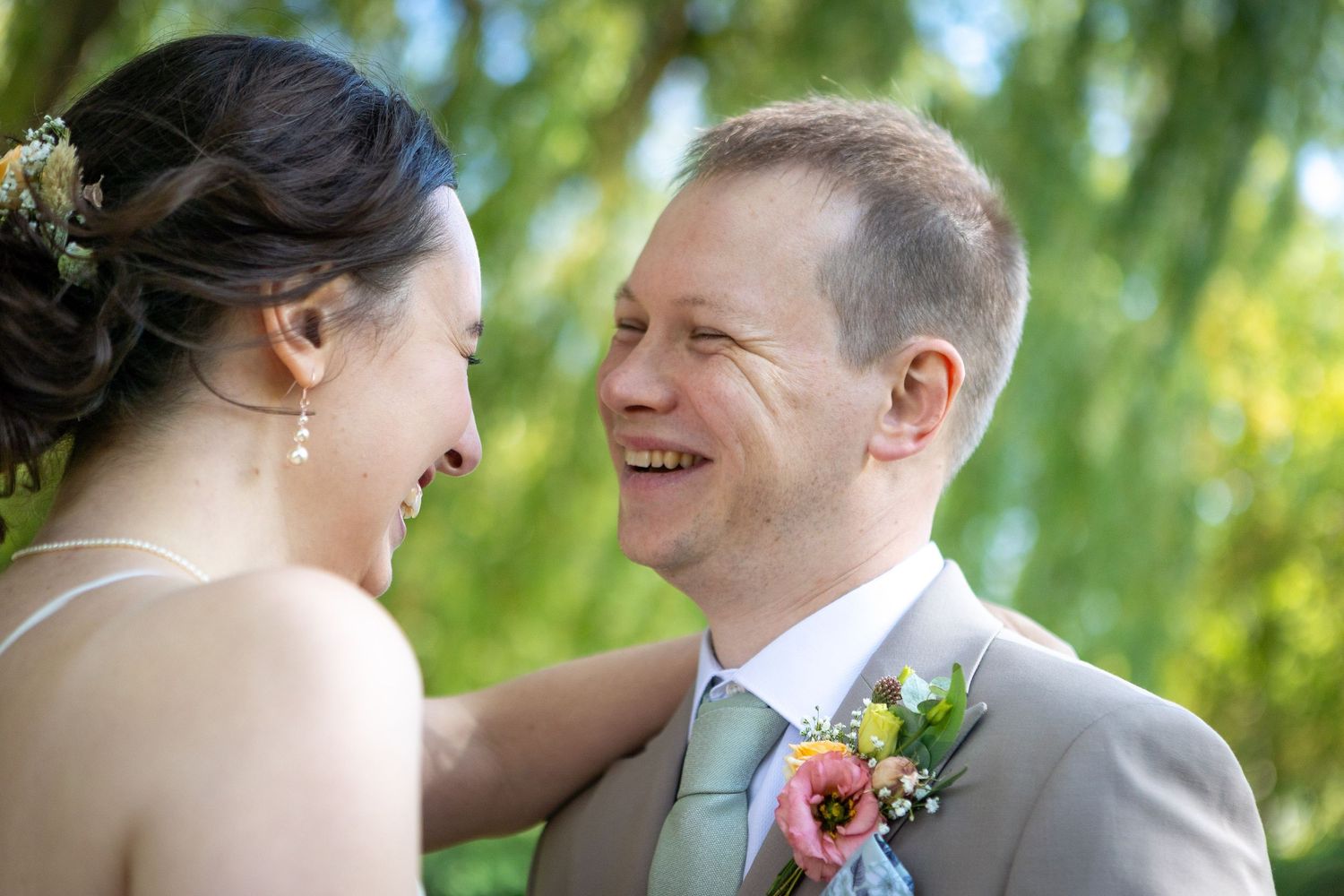 Groom laughing joyfully as bride touches his shoulder under green weeping willow branches.
