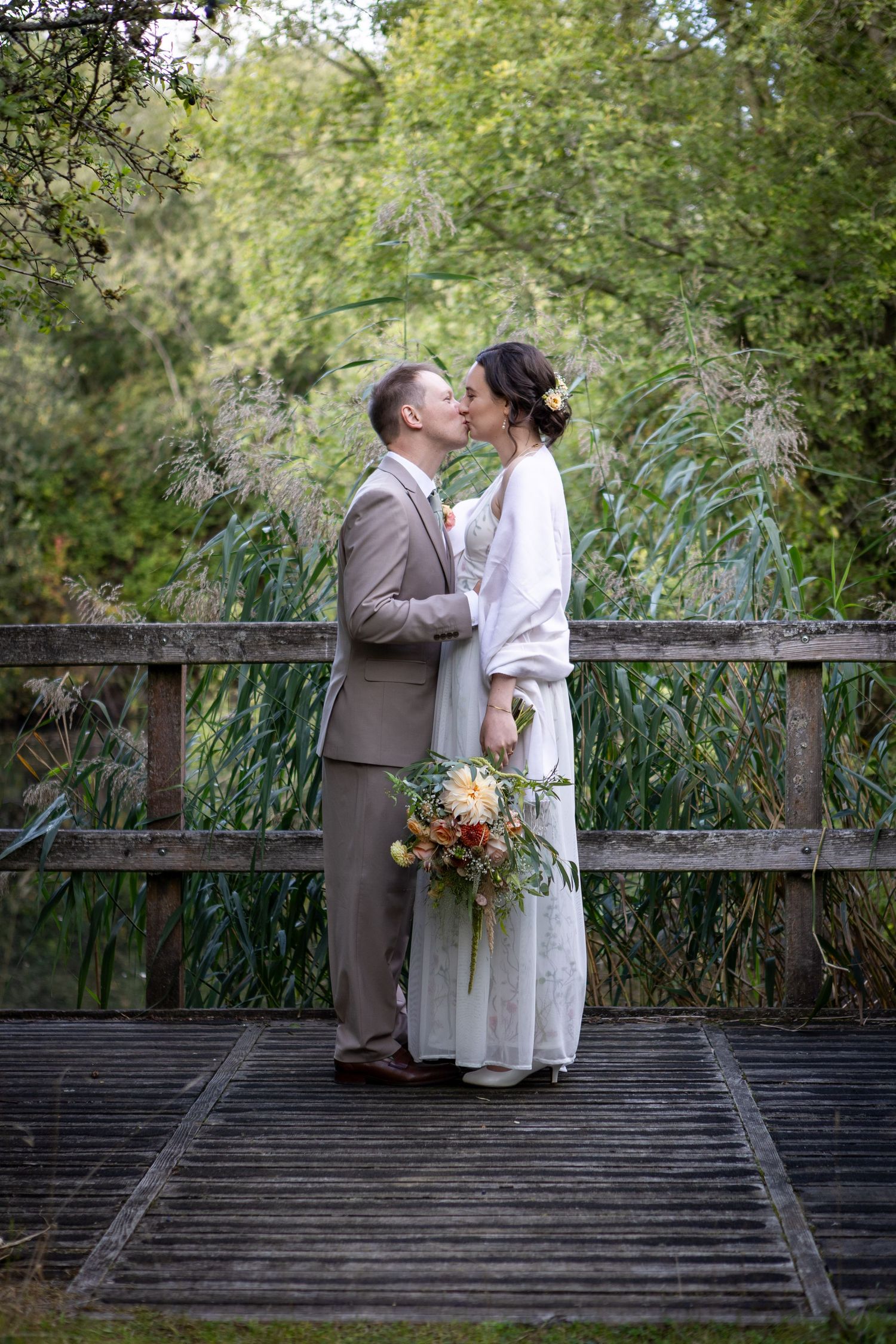 Bride and groom sharing a romantic kiss on a rustic wooden bridge surrounded by greenery.