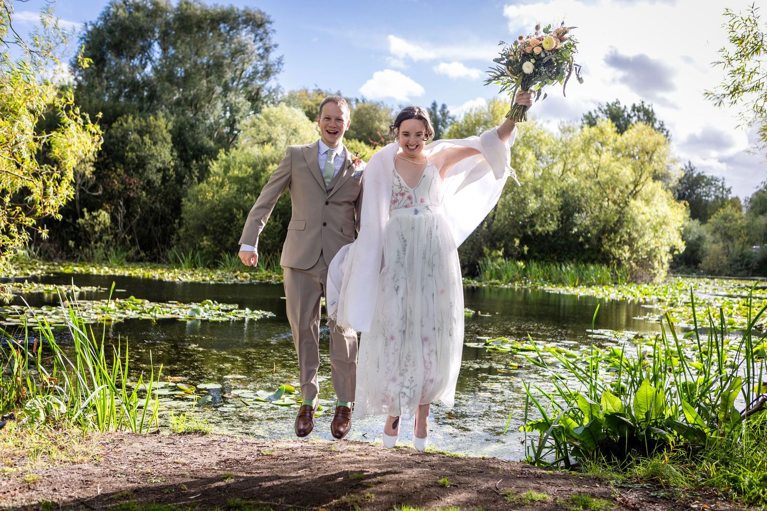 Joyful bride and groom splashing through a pond surrounded by lush greenery on their wedding day.