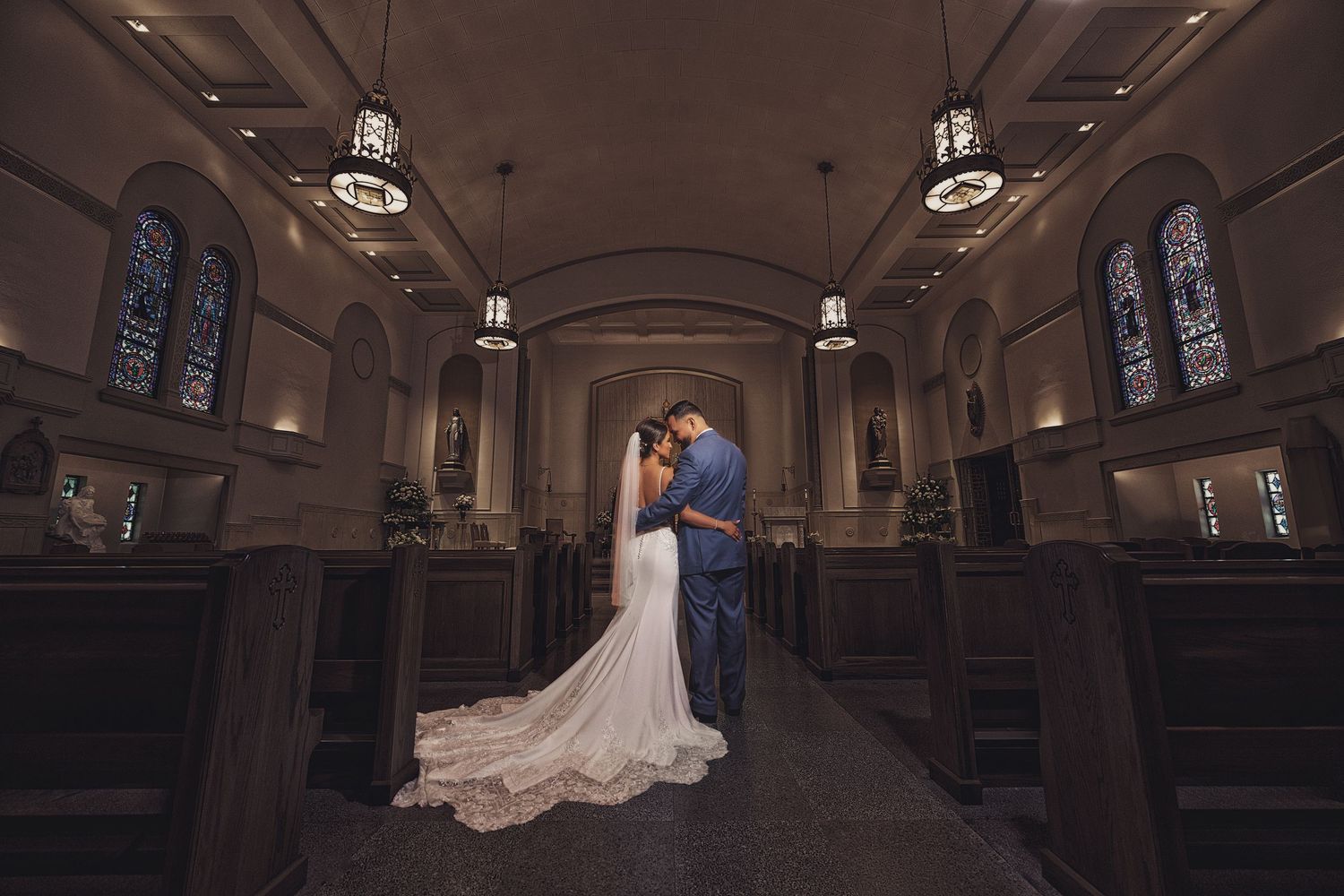 A bride and groom embrace in a dimly lit cathedral aisle, framed by wooden pews, hanging lanterns, and colorful stained glass windows.