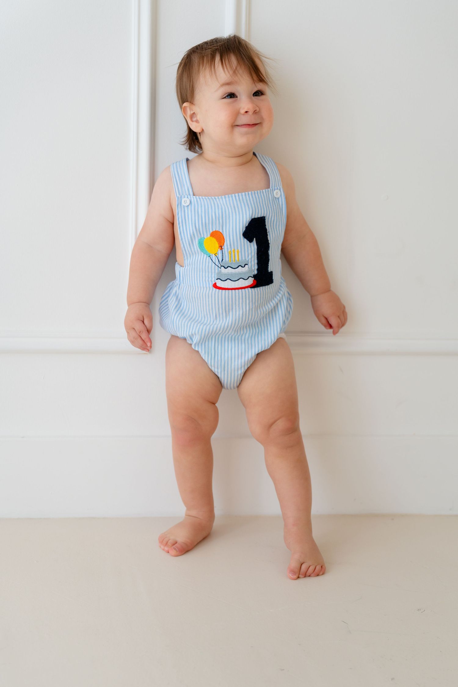 Baby boy in blue seersucker romper with birthday cake and number 1 applique, standing barefoot against white wall.