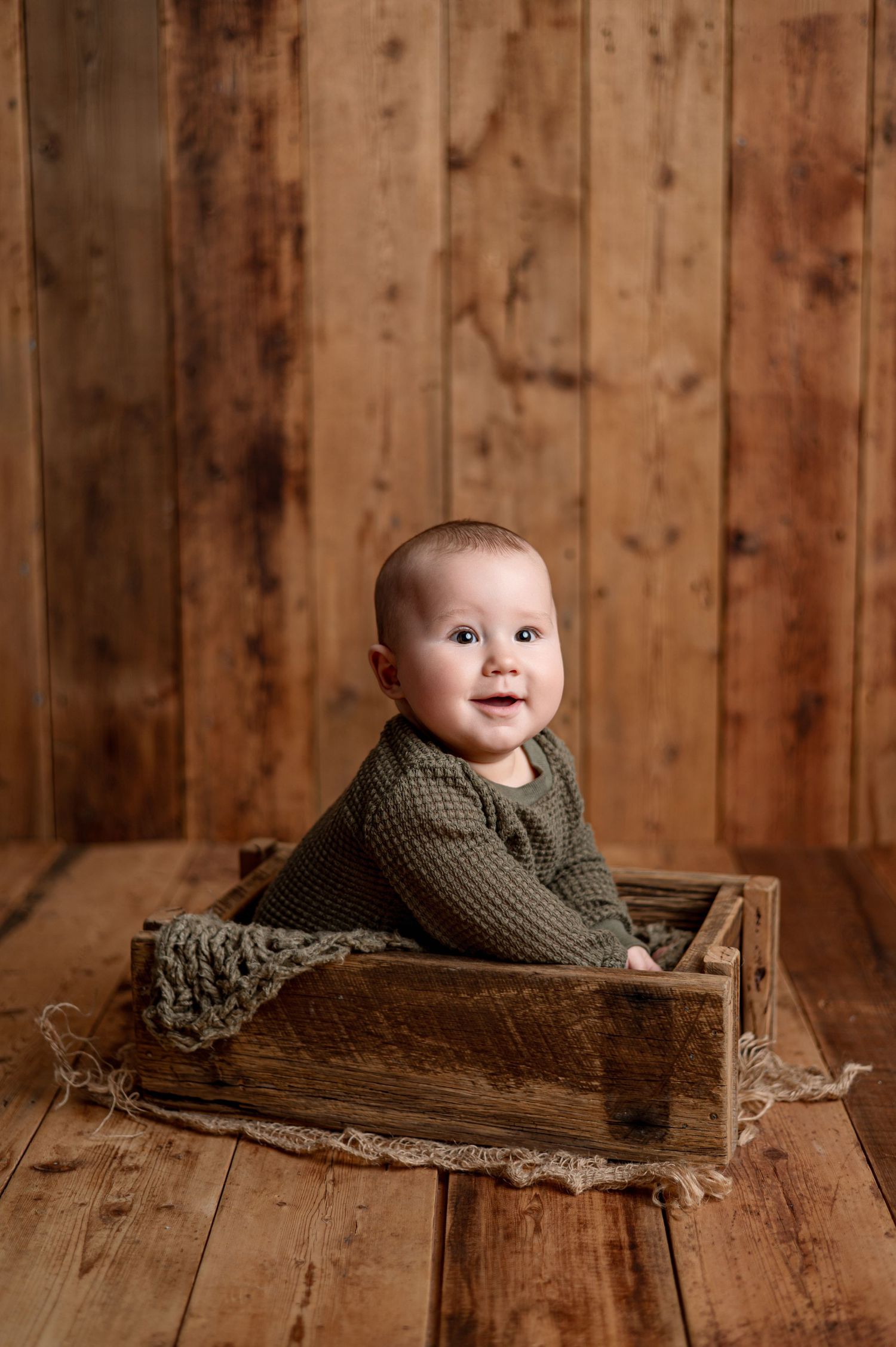Baby girl in yellow dress sits in wicker chair with floral headbow, purple backdrop with white flowers.