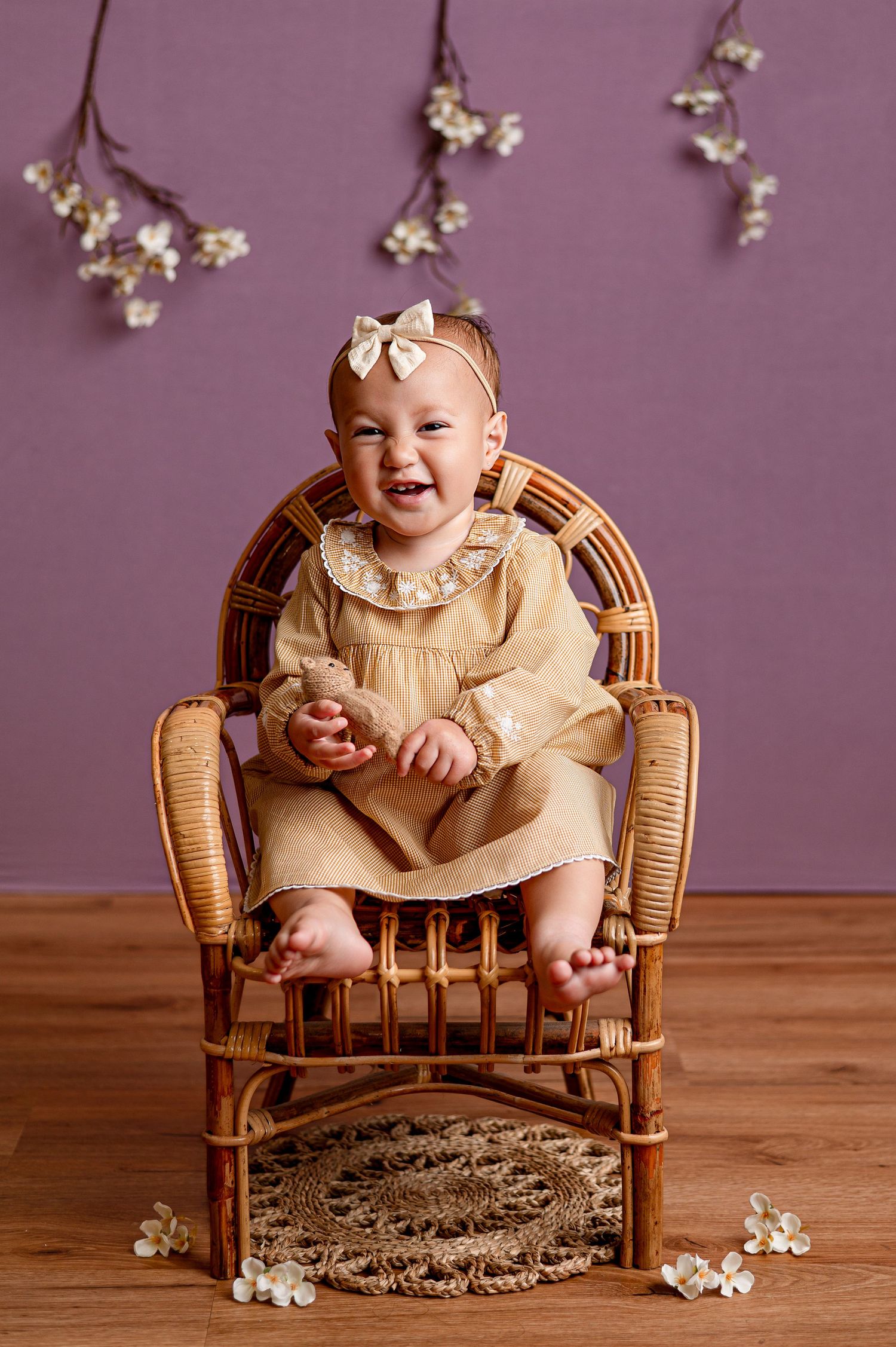 Baby girl in yellow dress sits in wicker chair with floral headbow, purple backdrop with white flowers.
