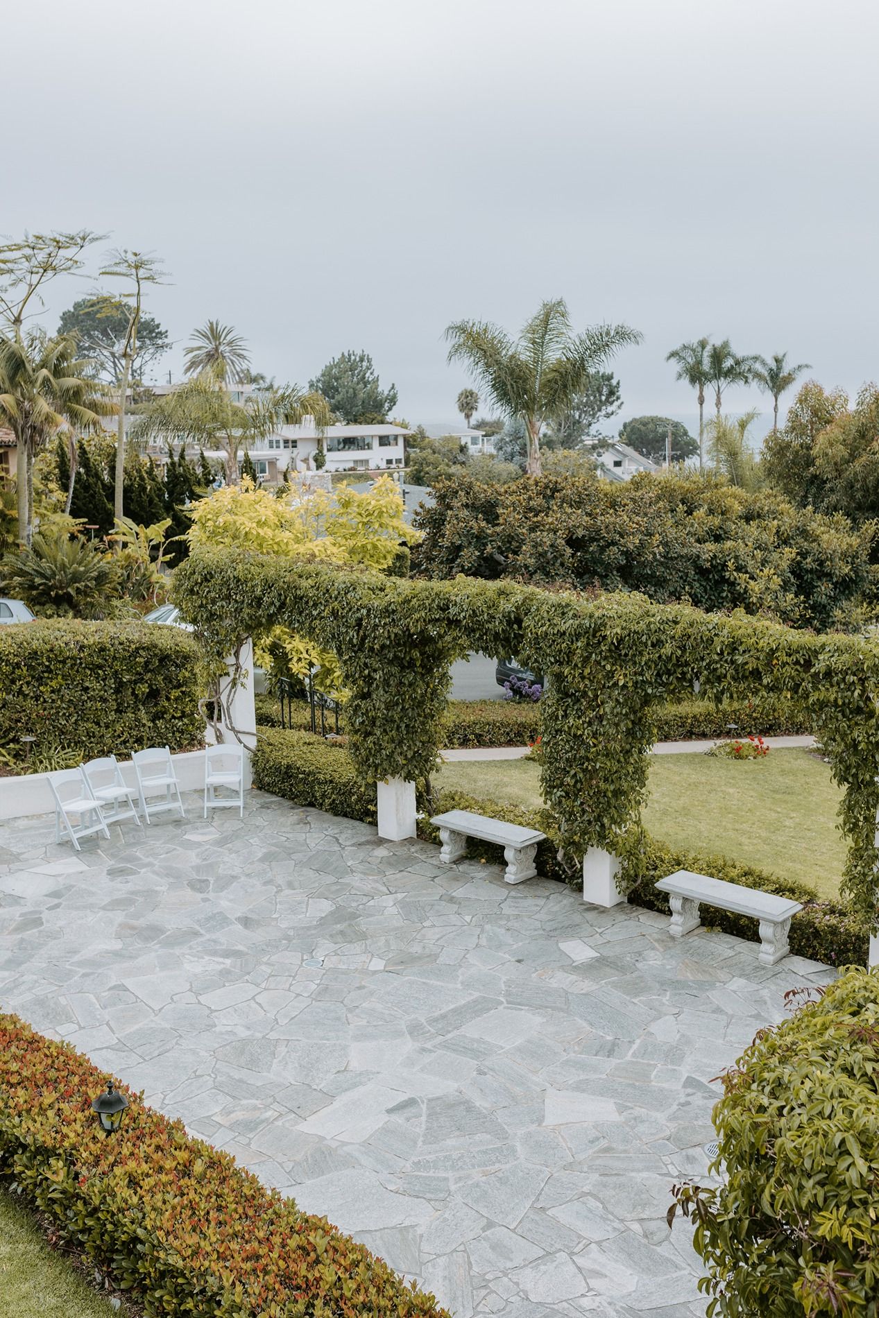 Manicured garden at The Thursday Club with trimmed hedges, stone pathways, white benches, and palm trees under an overcast sky.