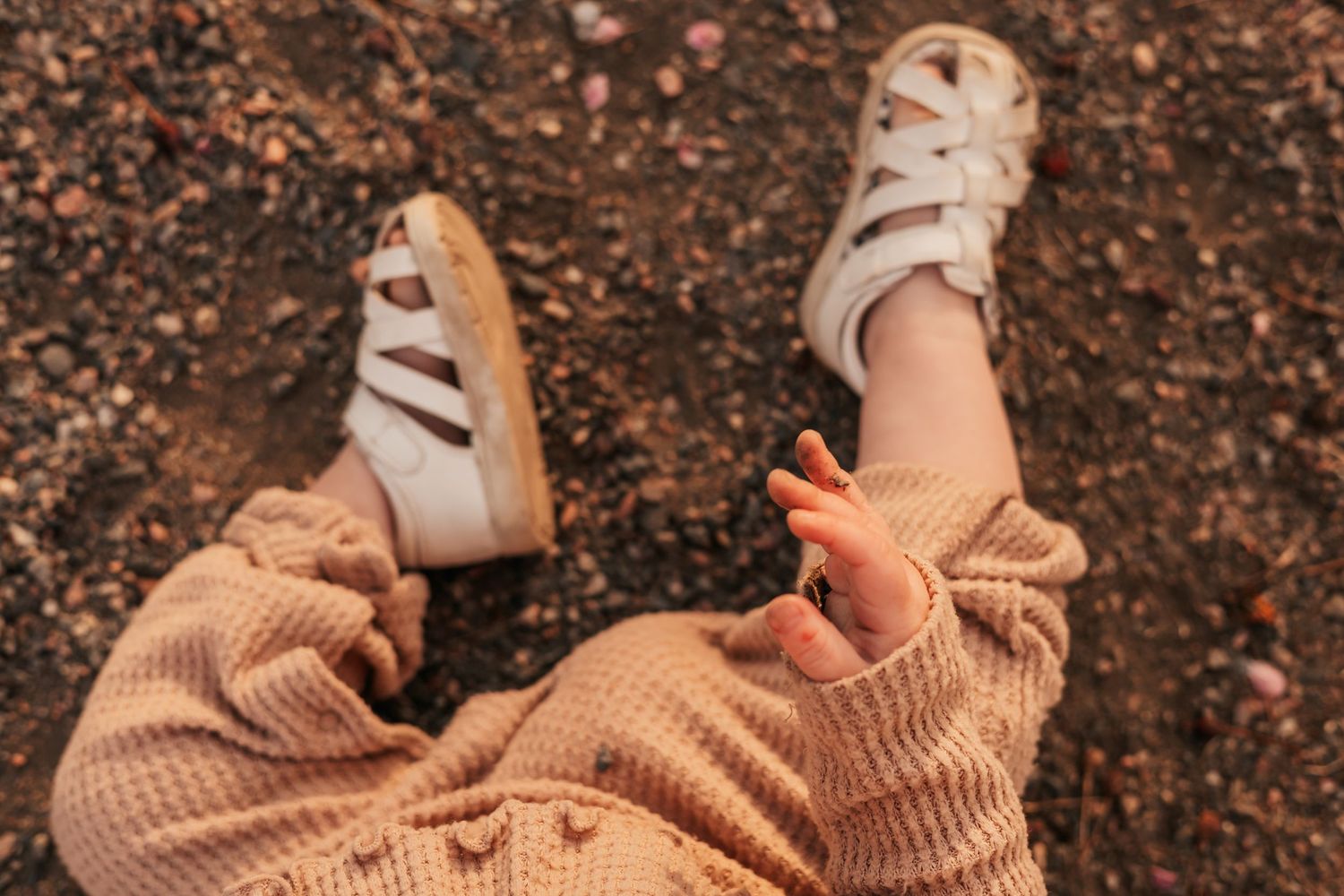 Close-up of a baby wearing white sandals and a beige waffle-knit outfit, sitting on a pebbly ground | How to Get Nice Family Photos with a W