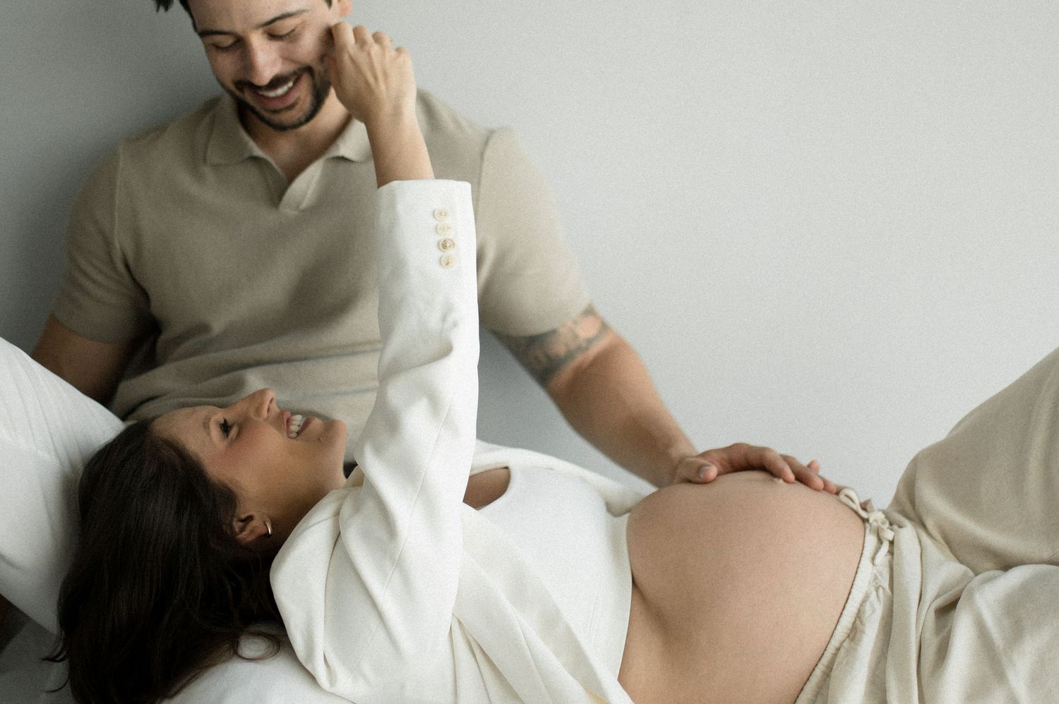 A pregnant woman in casual attire of white shirt and jeans poses against a neutral background in a maternity photo.