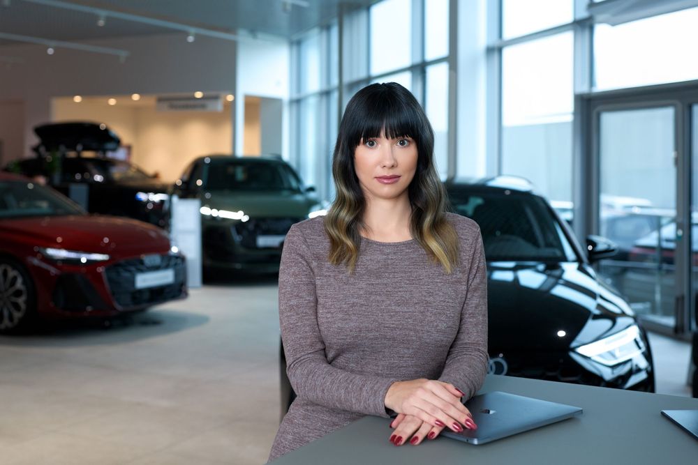 Woman in grey top leans on desk in modern car dealership showroom with luxury vehicles displayed behind her.