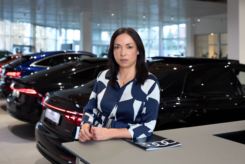 Professional woman in blue patterned blouse standing in luxury car dealership showroom surrounded by black vehicles.