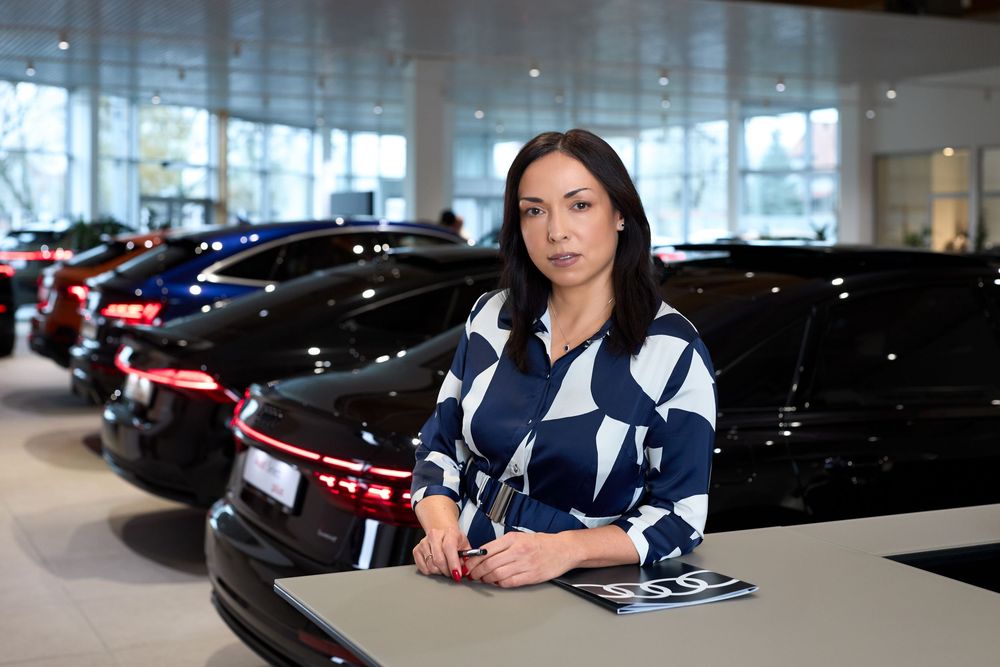 Professional woman in blue patterned blouse stands at desk in upscale car dealership showroom.