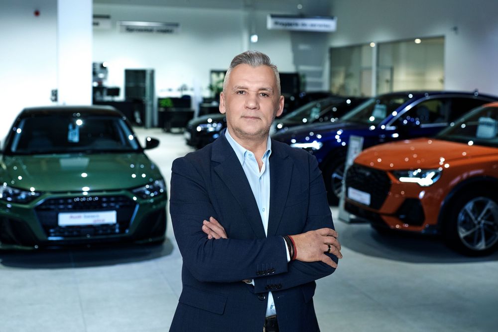 Man in blazer poses confidently in modern Audi dealership showroom with green and orange SUVs displayed.