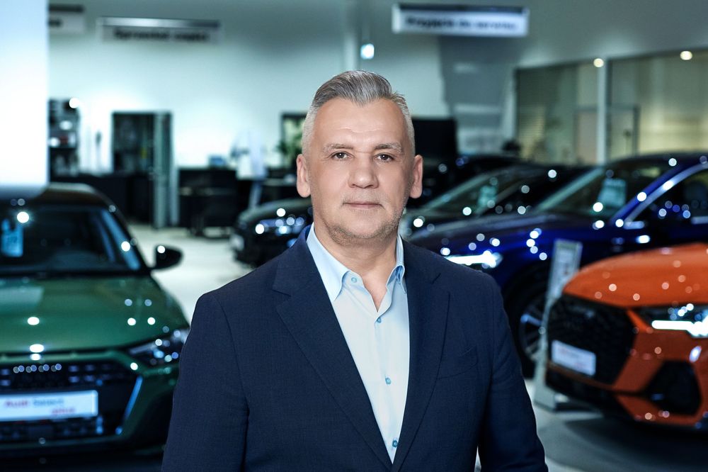 Middle-aged man in a suit standing in an Audi car dealership showroom with colorful vehicles displayed behind him.