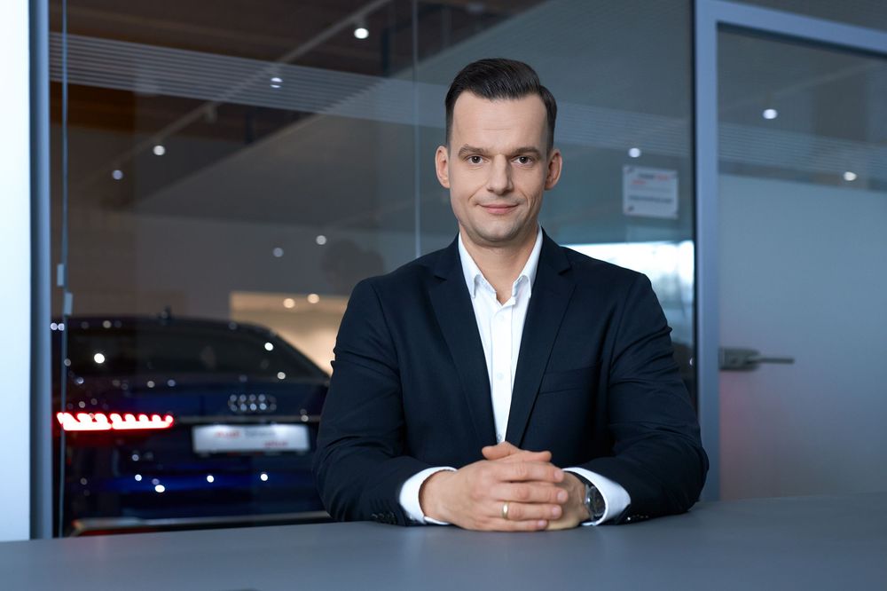 Professional businessman in dark suit sits at desk in modern Audi dealership showroom with blue lighting.