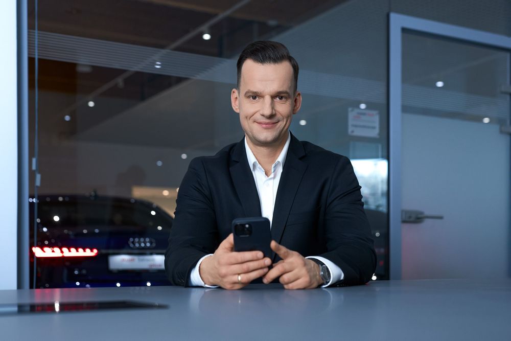 A professional businessman in a black suit holds a smartphone while sitting at a modern desk in an Audi showroom.