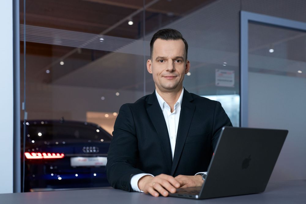 Professional businessman in dark blazer working on laptop in modern Audi car dealership showroom.