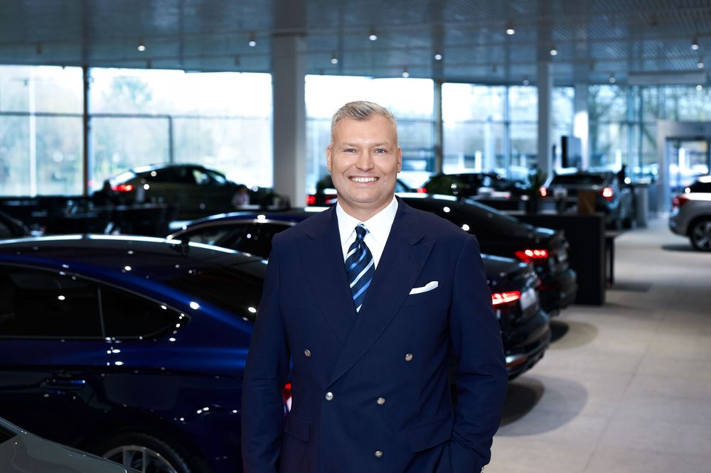Smiling businessman in navy suit stands in premium car dealership showroom with luxury vehicles behind him.