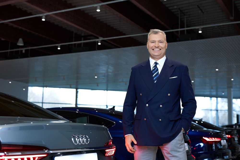 Smiling businessman in navy suit stands confidently among Audi vehicles in a modern car dealership showroom.