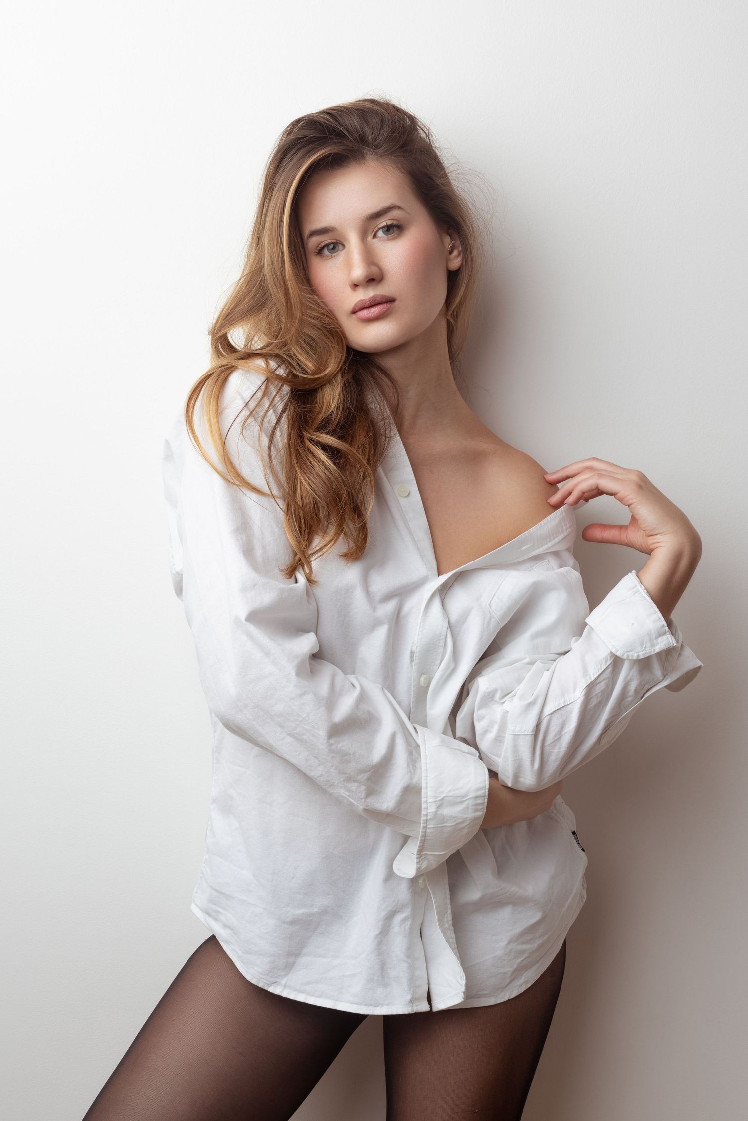 Young woman with long wavy hair posing in an oversized white button-up shirt and black tights against a white wall.