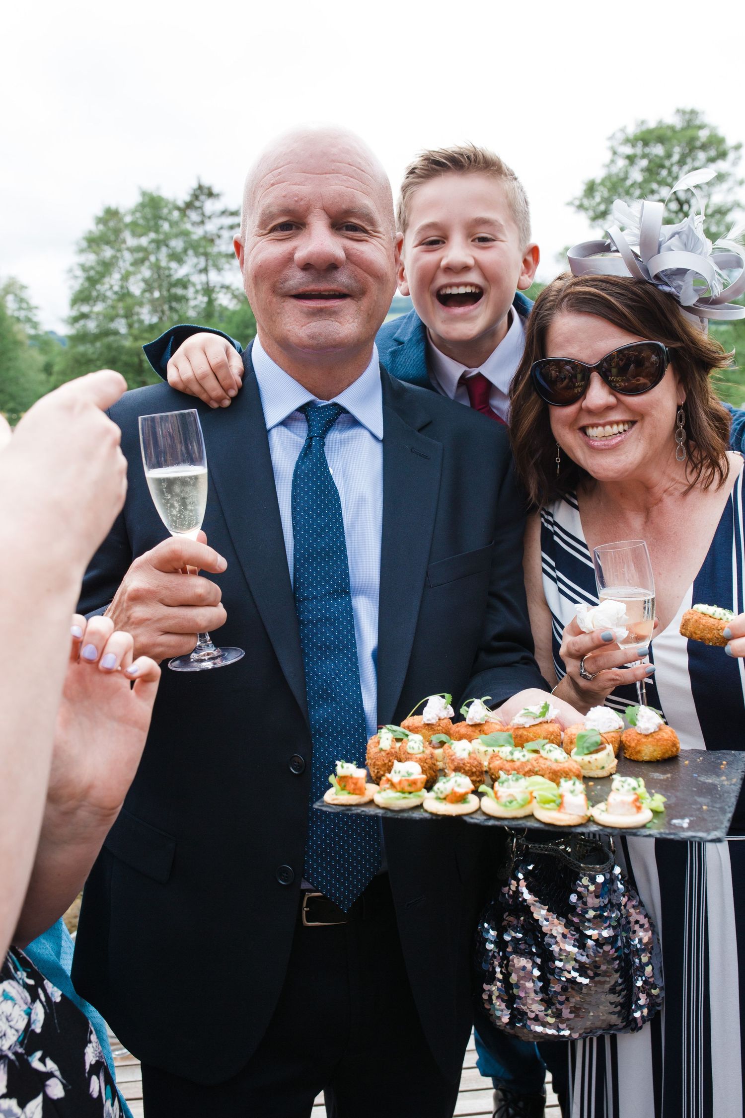 Family of three smiling at wedding reception holding champagne glasses and appetizer tray outdoors.