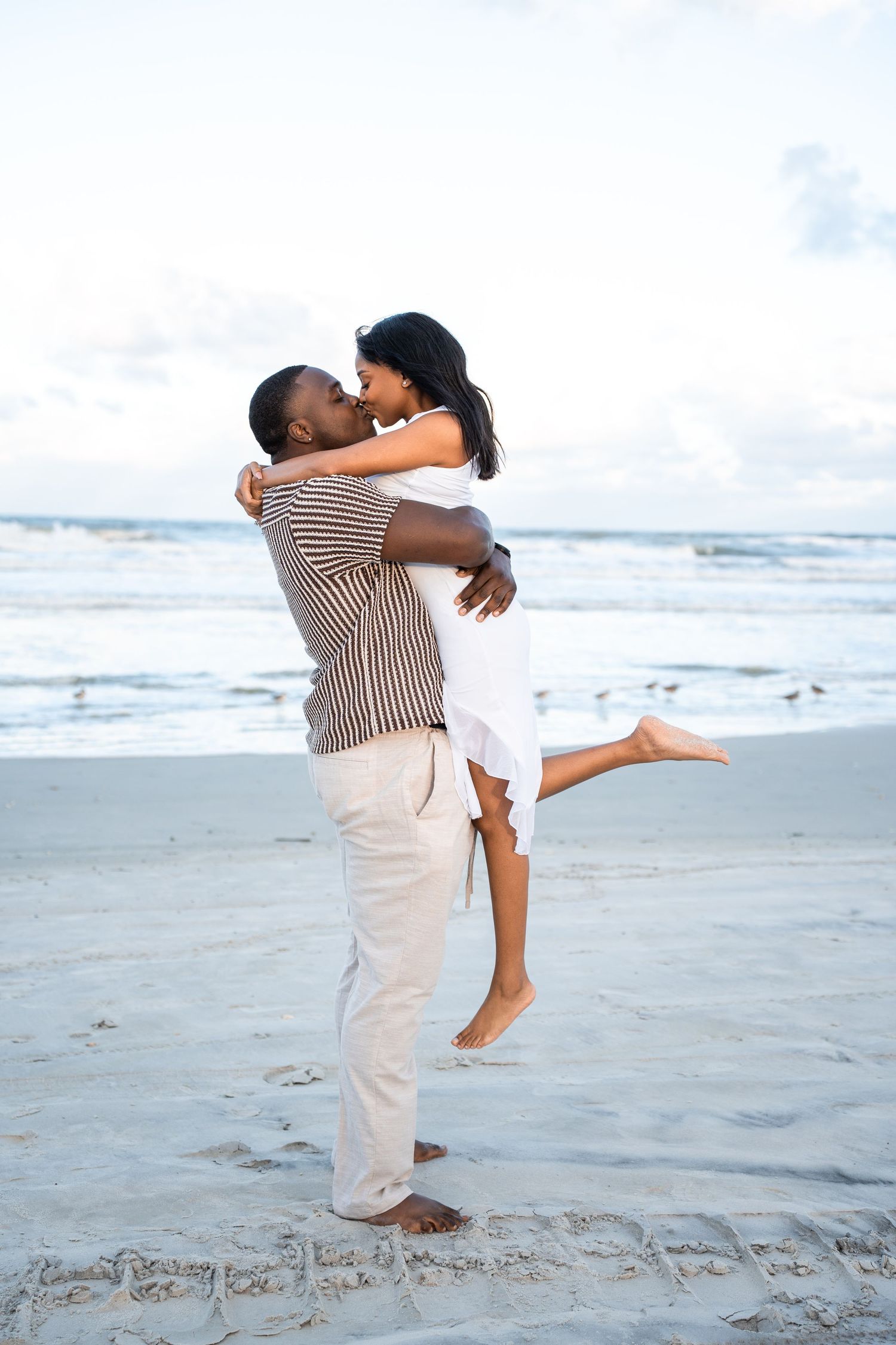 A romantic couple embraces and kisses on a sandy beach, with the man lifting the woman as ocean waves roll in behind them.